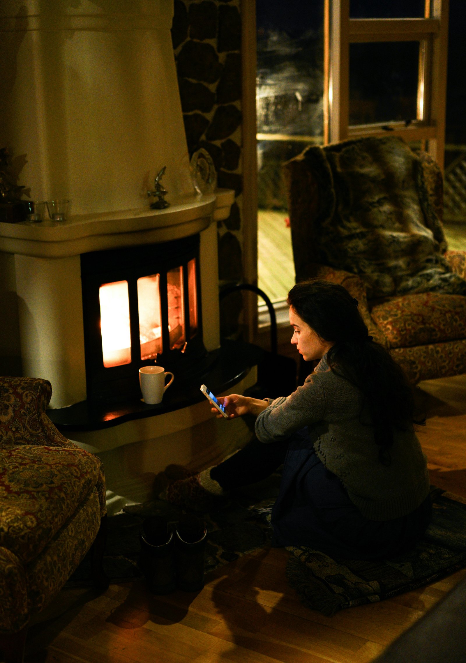 A young woman sits by a lit fireplace